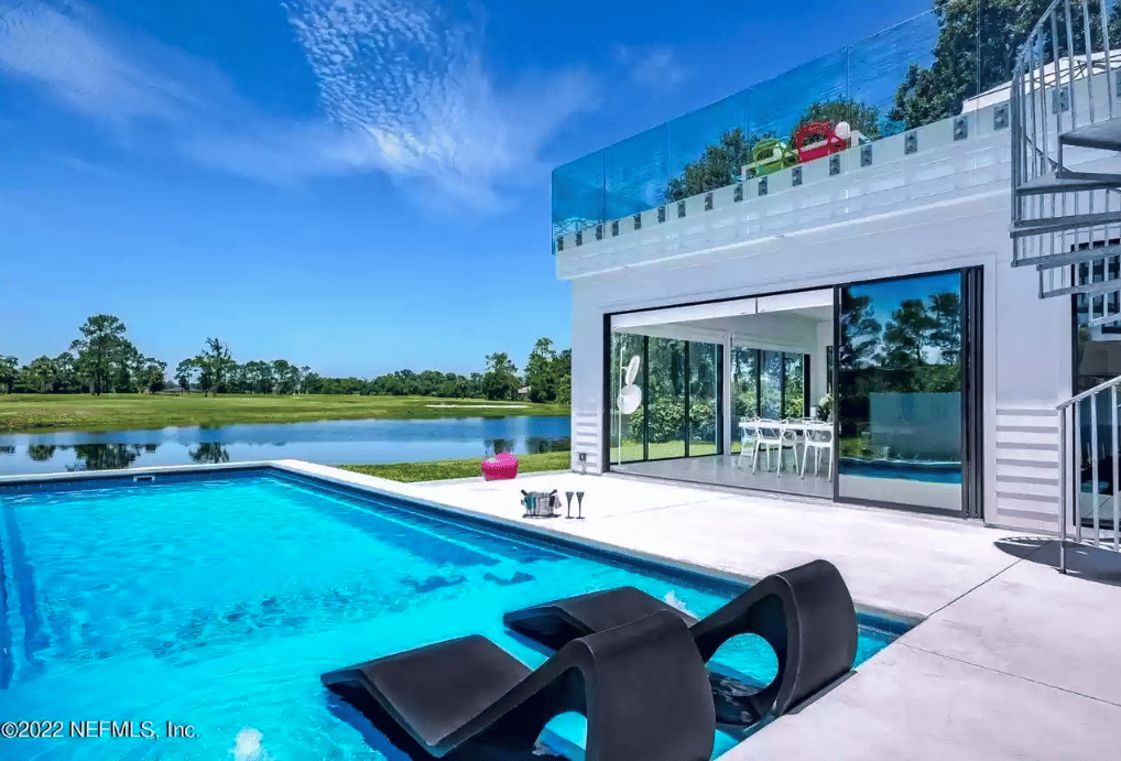 Modern home with a swimming pool and black loungers beside a pond.