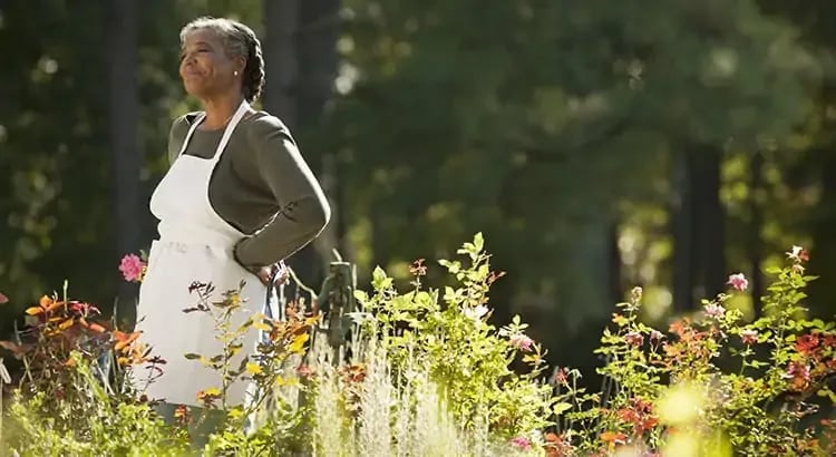 A woman in a garden, possibly harvesting or tending to plants. She is wearing a white apron, and the background is lush and green, suggesting a serene outdoor activity.