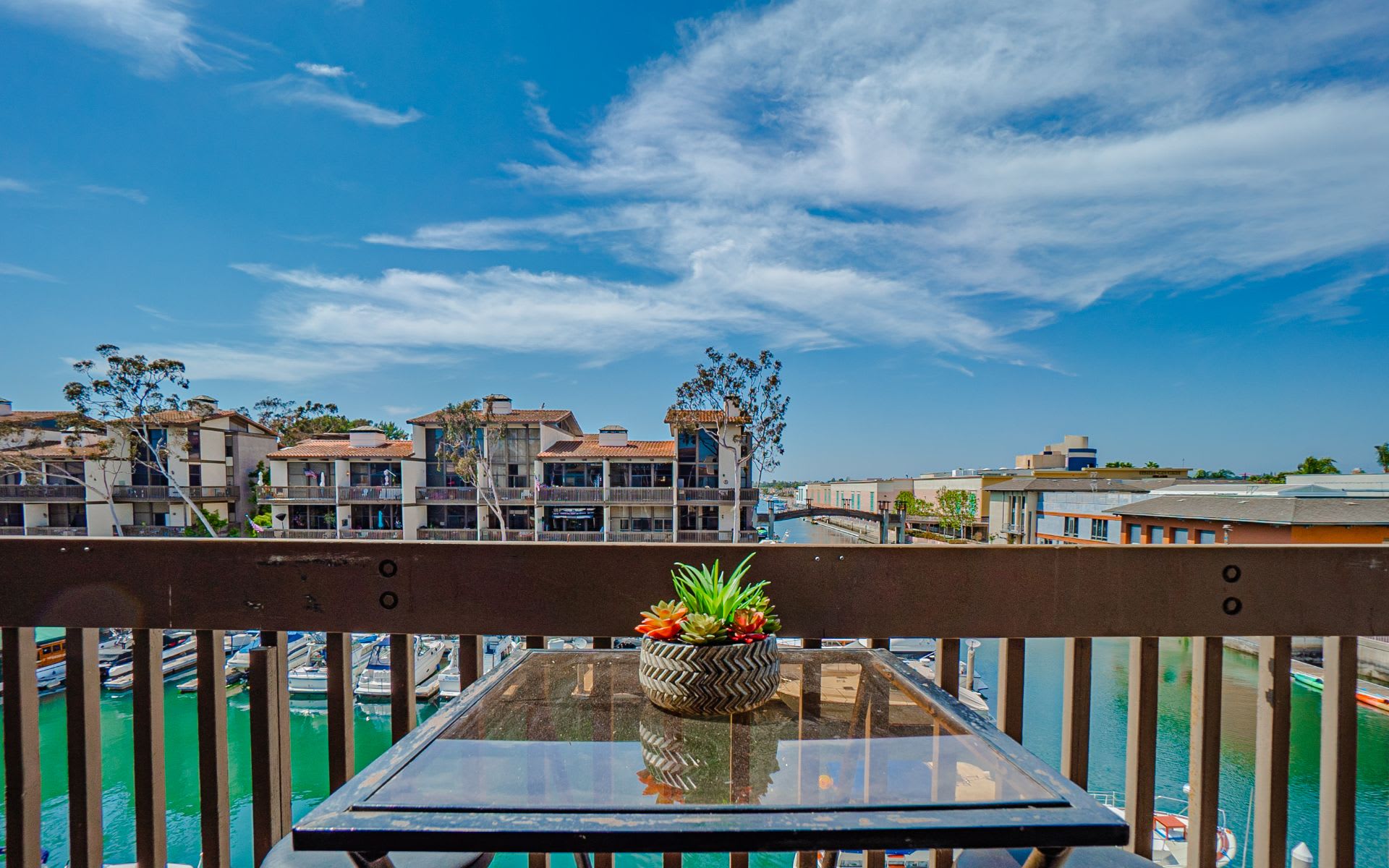 Condo balcony with a glass-top table overlooking marina water and neighboring waterfront buildings.