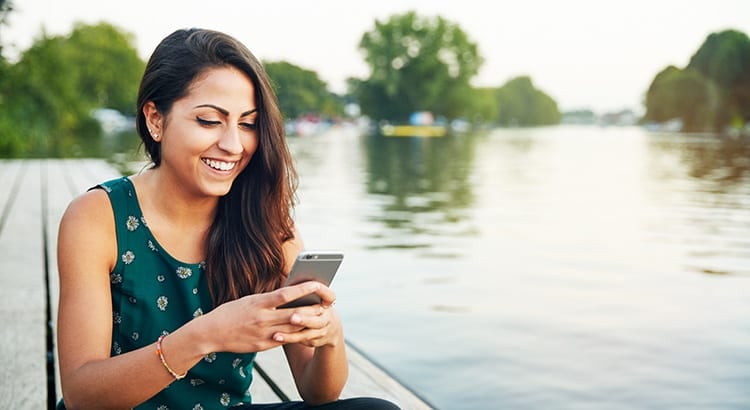 A woman smiling and using her smartphone while sitting by a body of water, suggesting themes of connectivity, lifestyle, or perhaps remote work.