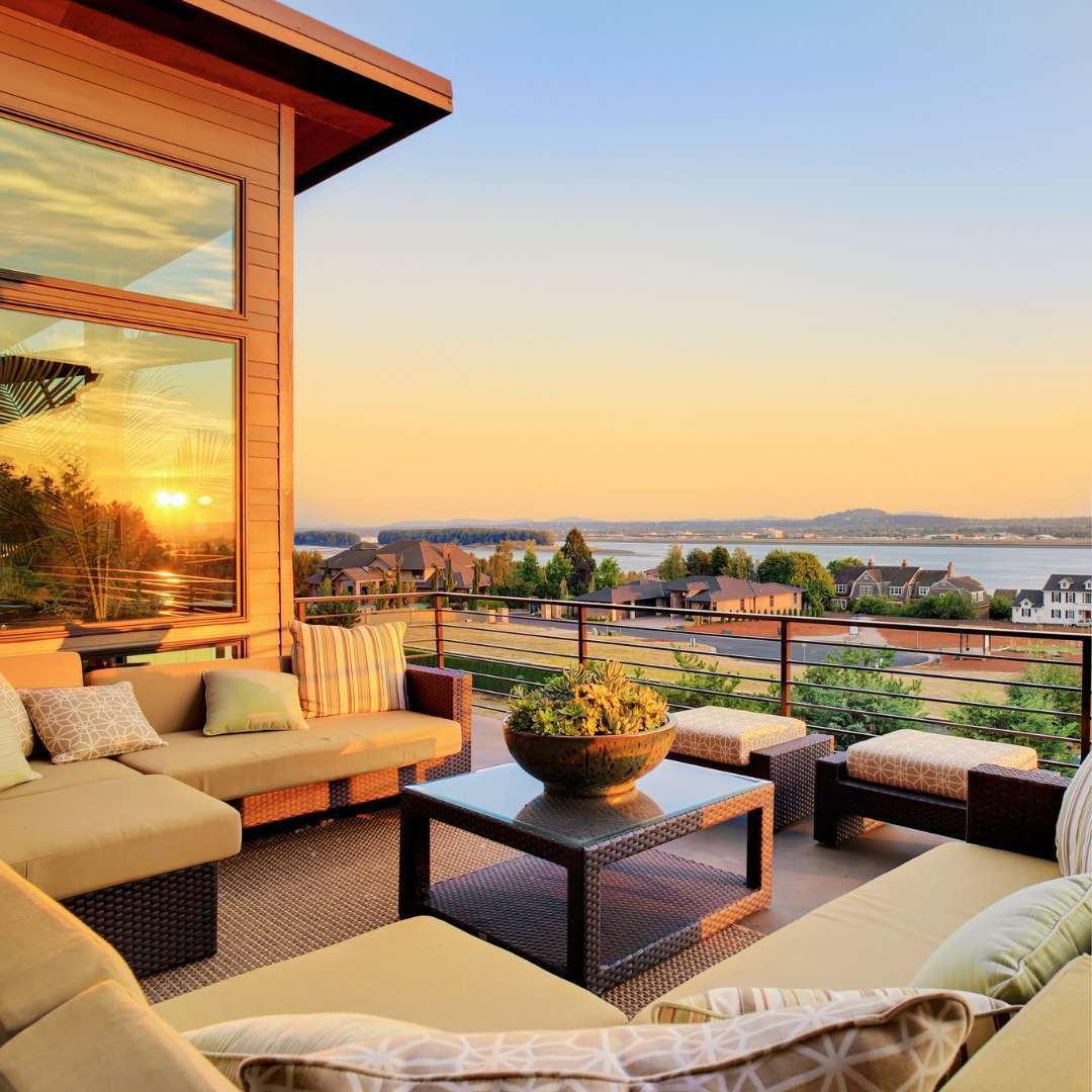 An inviting outdoor patio or balcony furnished with a sectional sofa and a coffee table, overlooking a scenic view of a body of water, distant land, and houses