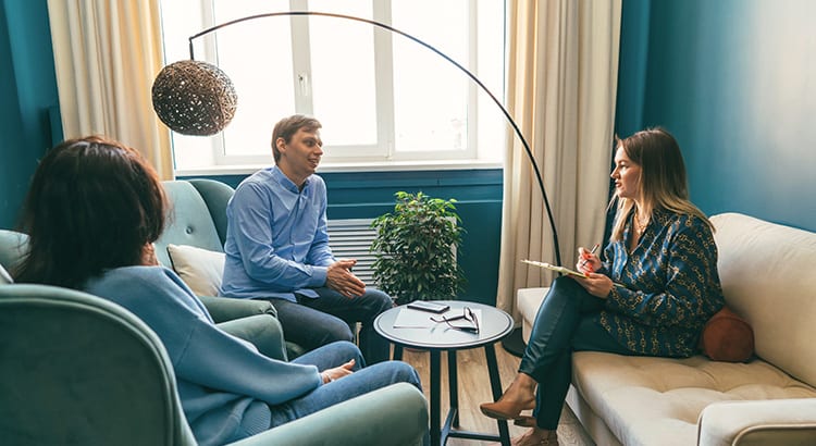 Three people having a discussion in a living room setting. The room has a cozy atmosphere with modern furniture and a large window with curtains.