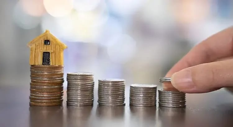 A stack of coins with a small house model on top and a hand placing another coin on the stack. This represents saving money, financial planning for home ownership, or investments.