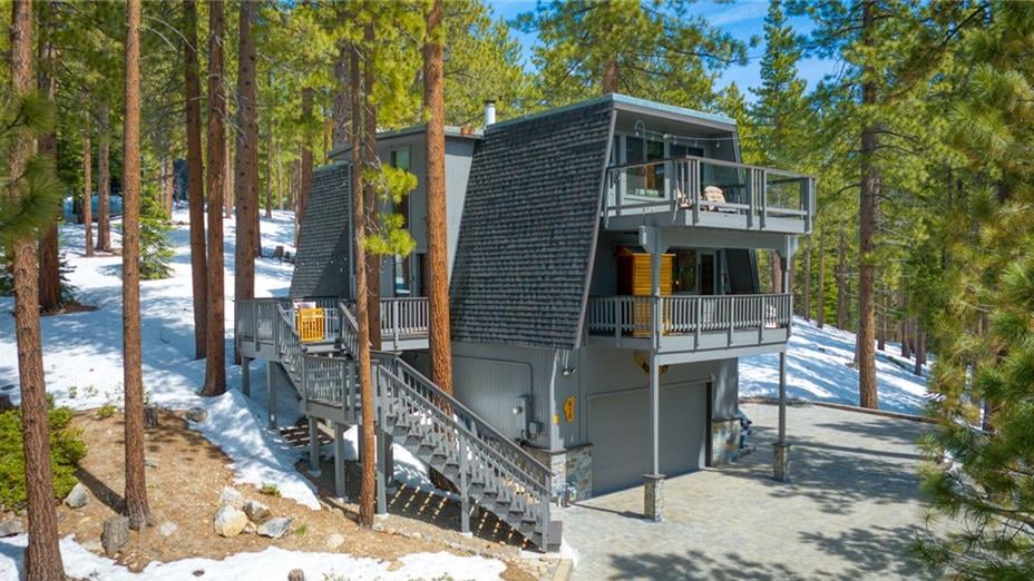 Aerial view of a gray, steep-roofed home with stacked decks, an exterior stair and a paver driveway among tall pines and spring snow at 808 Randall Avenue, Jennifer, Incline Village