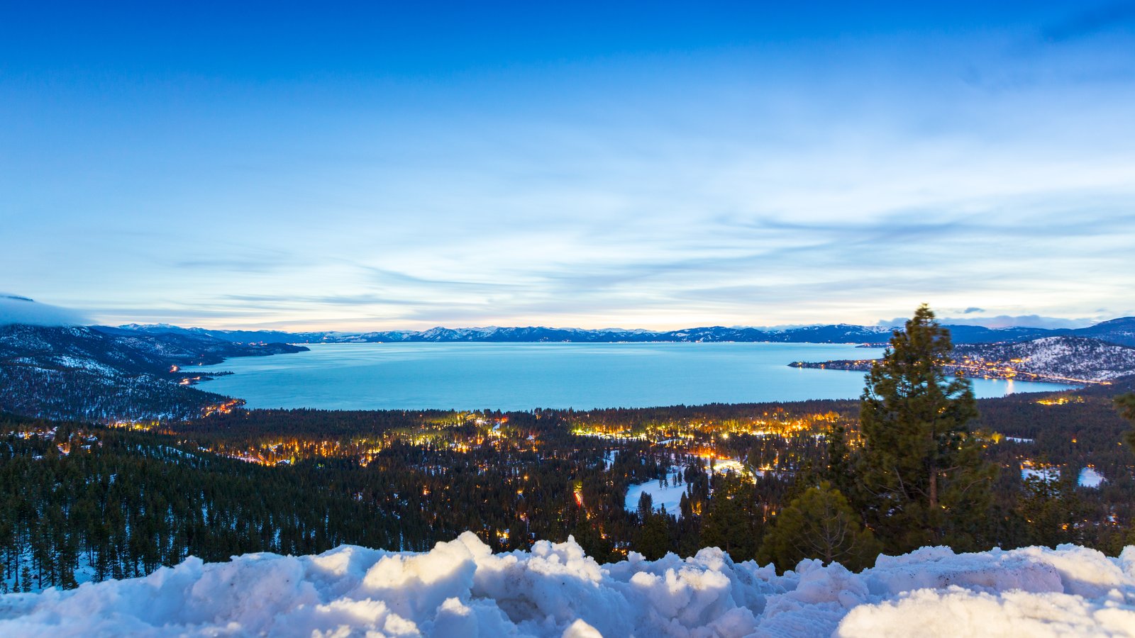 Lights of Incline Village glowing among snowy pines at dusk below the Mt. Rose Highway overlook, with Lake Tahoe and snowcapped peaks beyond