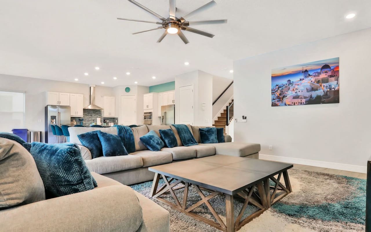 A living room interior with a couch, coffee table, ceiling fan, and a picture of Santorini on the wall.