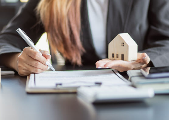 A person writing on a paper while holding a wooden house on the other hand.