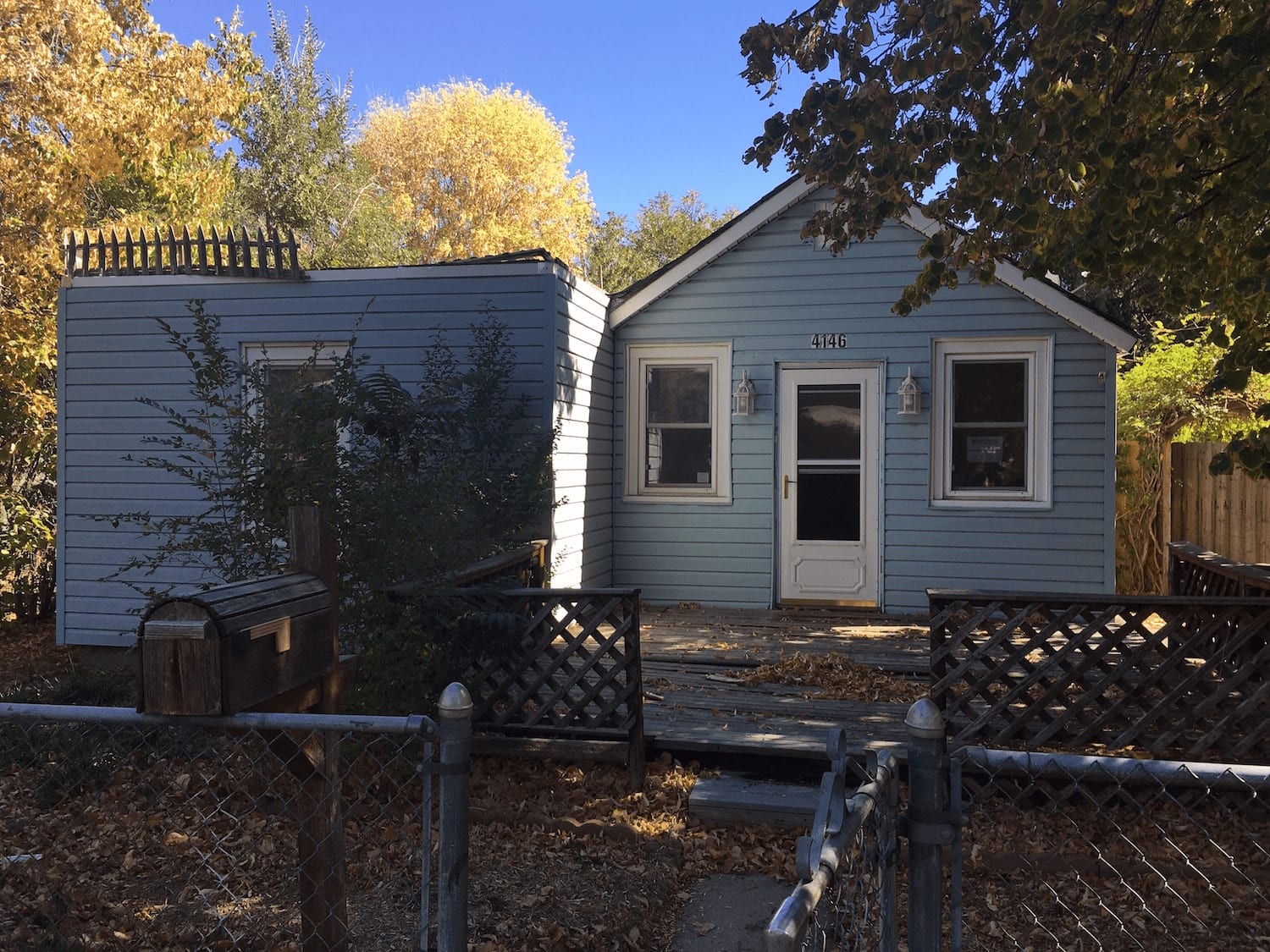  A single-story, light blue house with white trim and a white front door, featuring a small wooden porch area with lattice skirting.