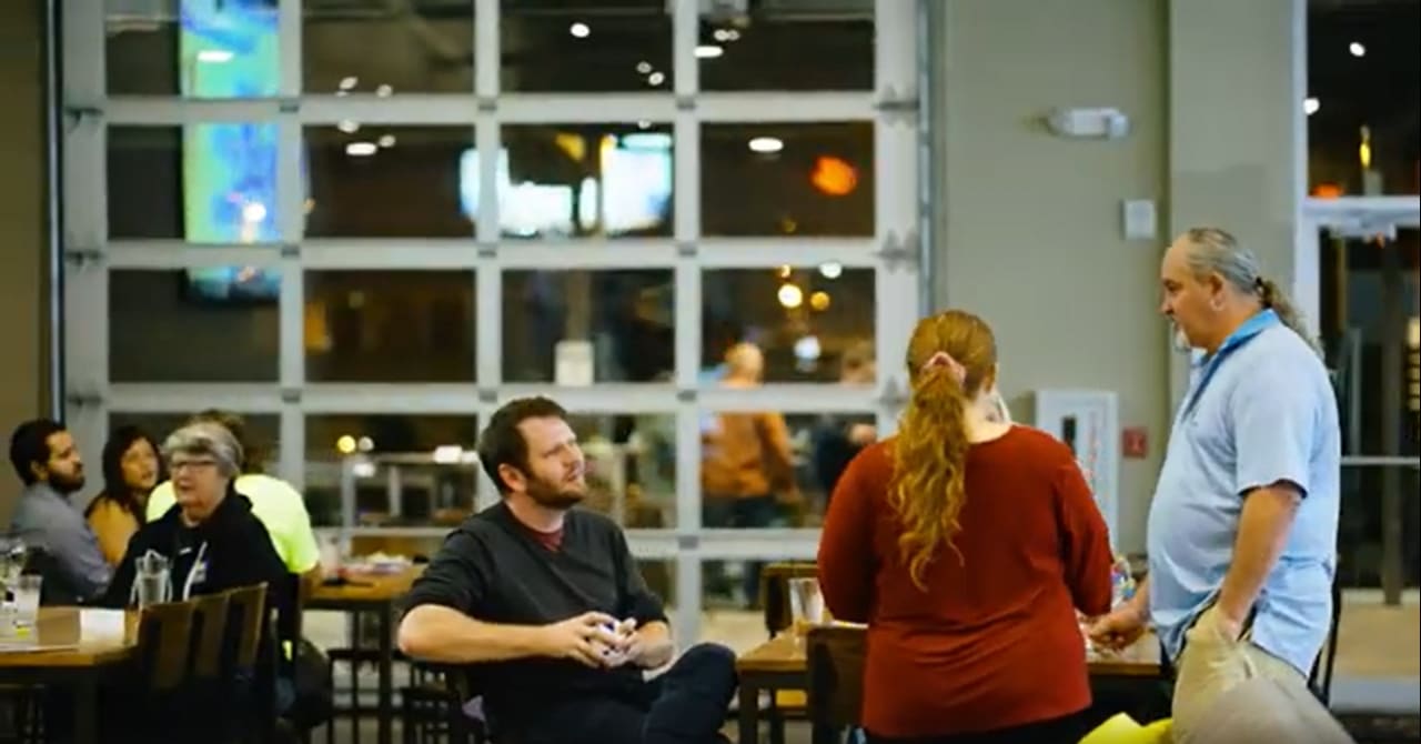 A group of people engaging in conversation inside a social setting, possibly a bar or cafe. The atmosphere is lively, with people seated and standing around.