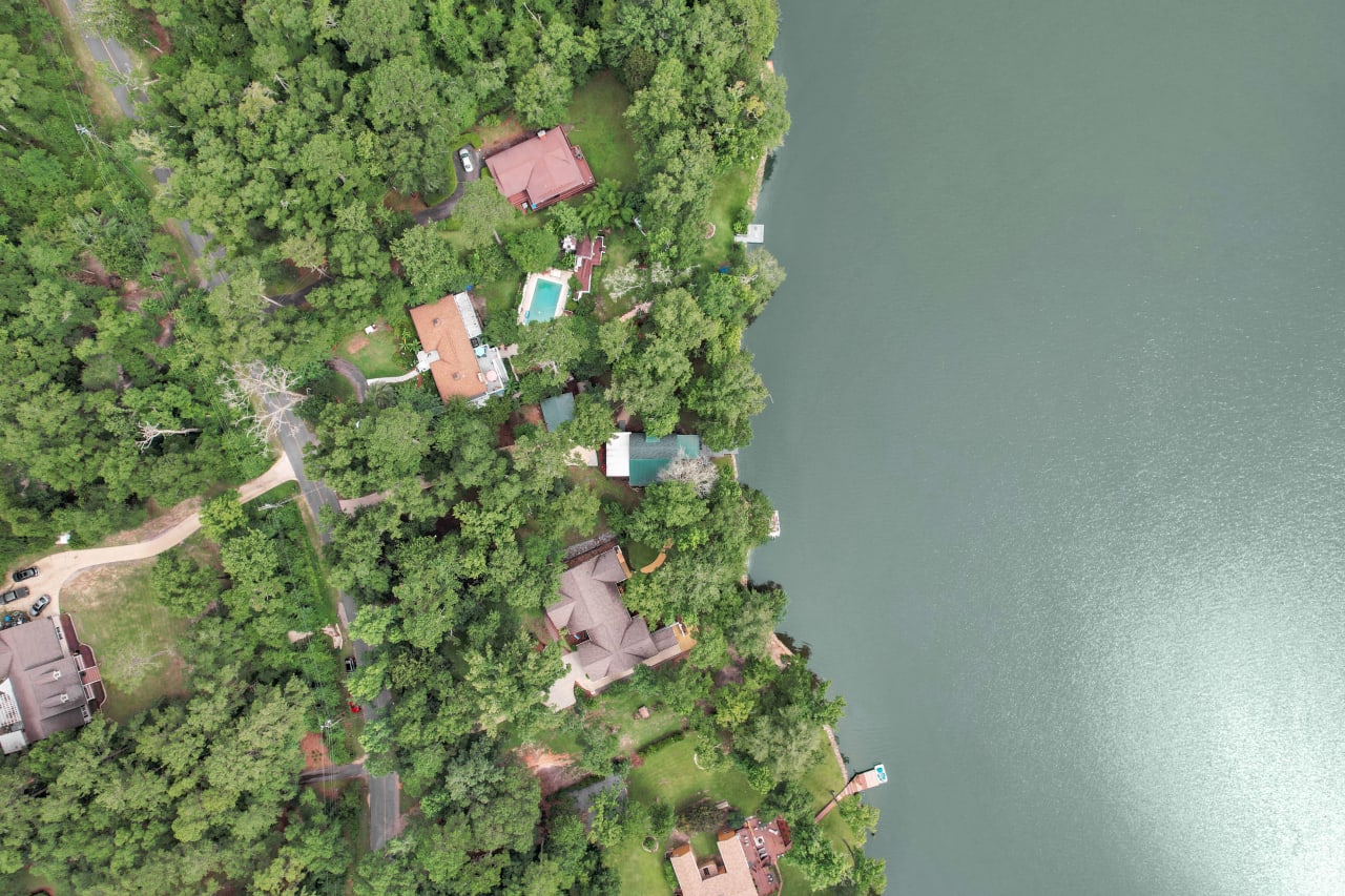 An aerial view of a green, forested area next to a body of water. Several buildings or houses are visible among the trees.