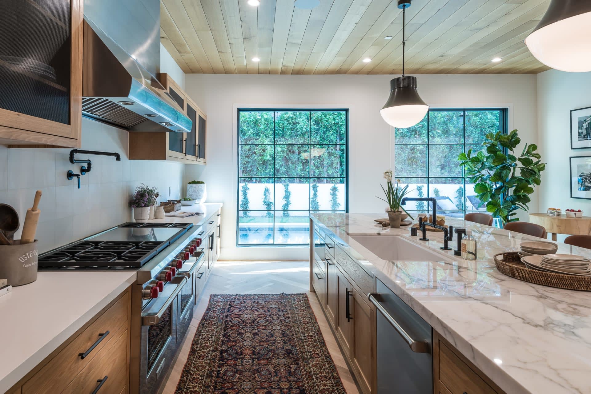 Modern kitchen with a marble island, wood cabinetry, gas range, and tall windows overlooking a pool.