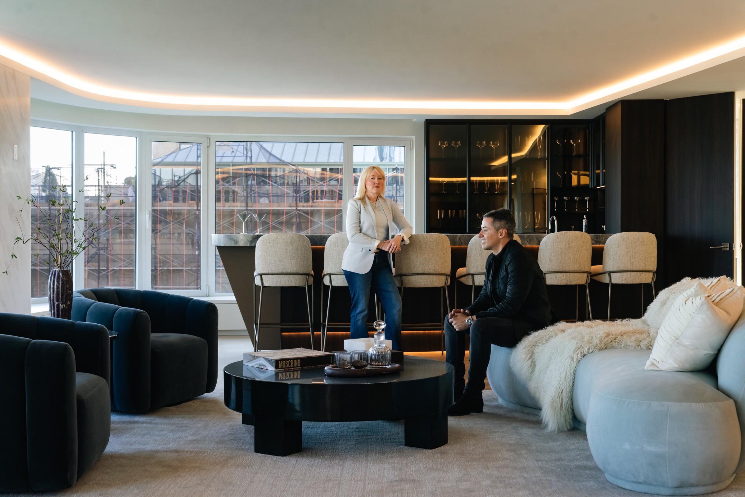 Brandon seated on the sofa looking toward Mallory beside the illuminated bar at 200 Central Park South