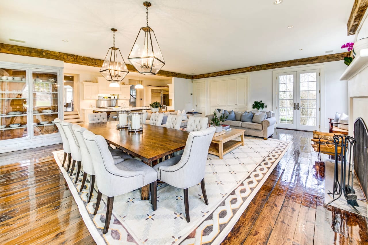 A dining room with a long table, chairs, fireplace, couch, and a door offering views outside.