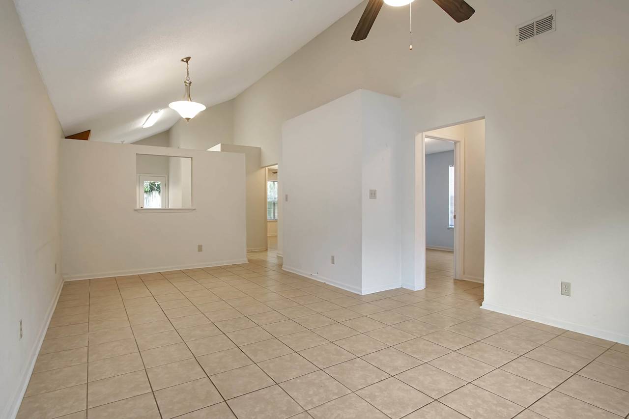 Photo of the main living area featuring vaulted ceilings, White walls, lightly colored tiles, a ceiling fan with light fixture, a pendant light over the dining area, a pass through window to the kitchen, the hallway to the guest bathroom and additional bedrooms, and the hallway to the primary bedroom  at 2709 Oak Park Court, Tallahassee, Florida 32308