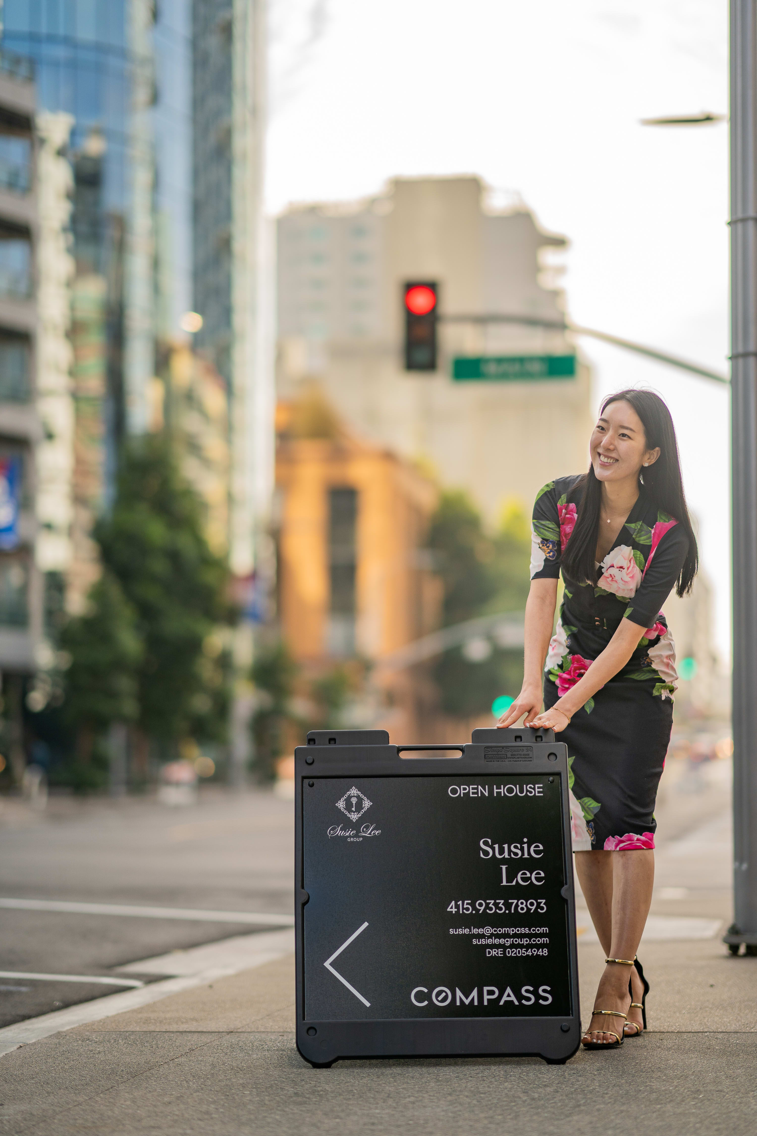 Woman in a black floral dress beside a Susie Lee Group open house sign on a city sidewalk.