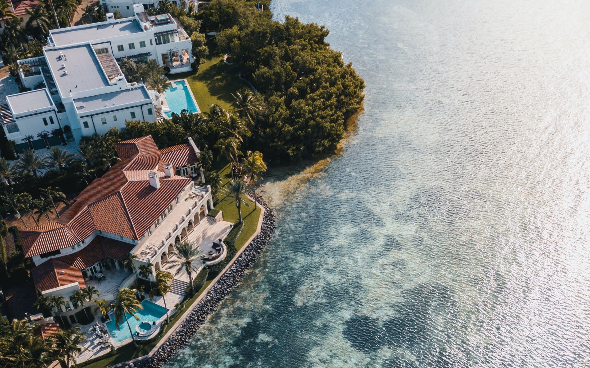 Aerial view of waterfront homes with red tile roofs, palm trees, and swimming pools.
