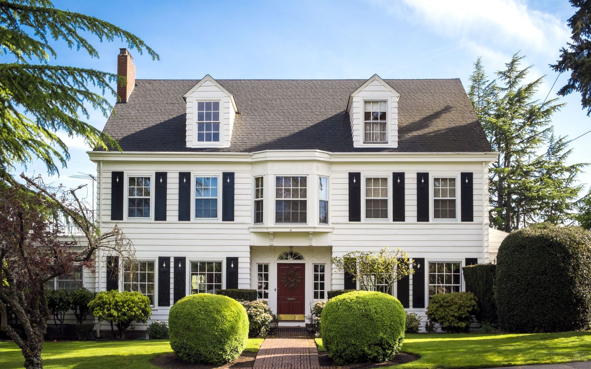  A grand, two-story white colonial-style house with a dark roof stands prominently, flanked by lush green trees under a clear sky.