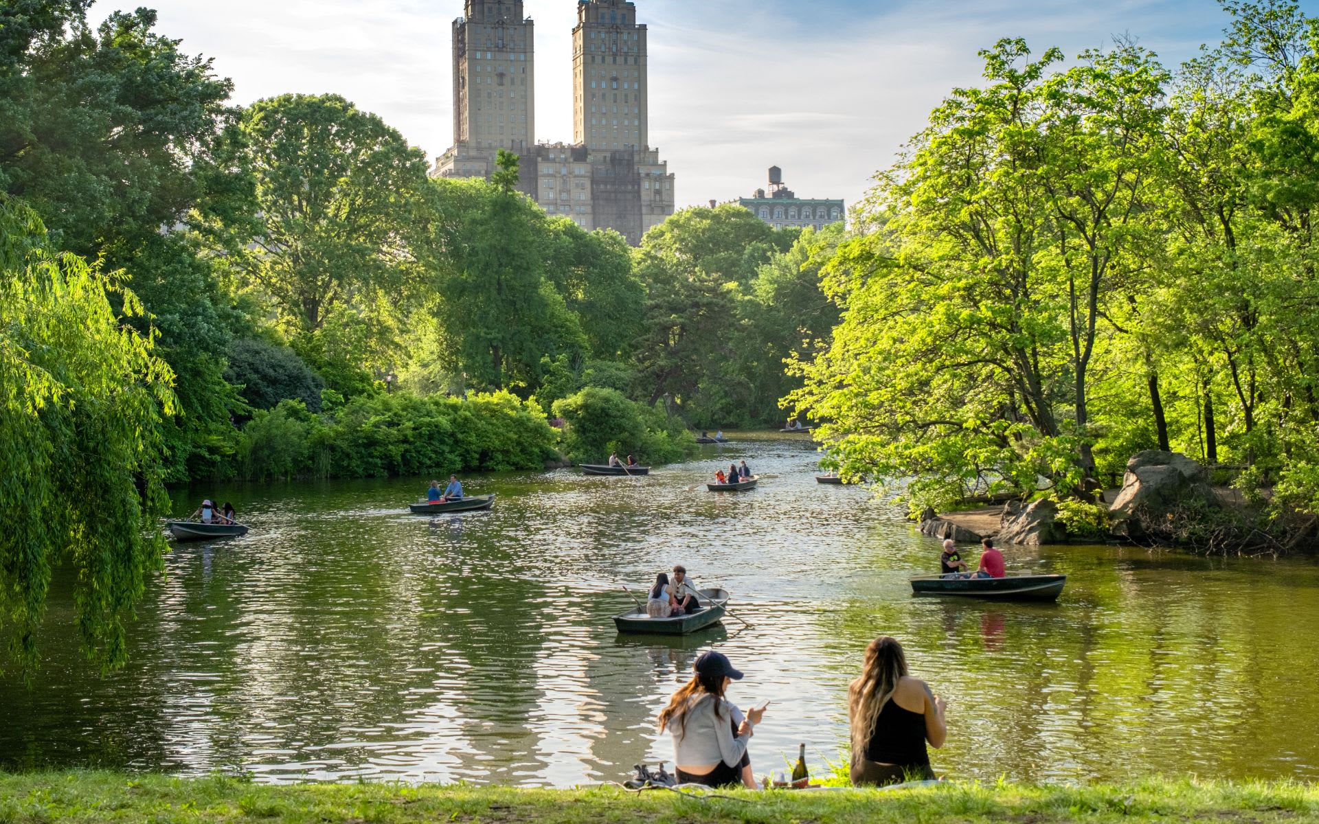 Rowboats on a Central Park lake beneath leafy trees and Manhattan apartment towers.