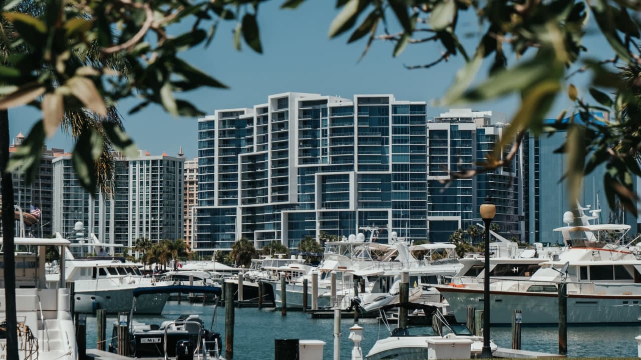 Modern high-rise buildings line a bustling marina filled with yachts and boats, framed by lush foliage.