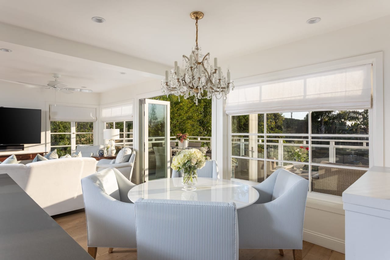 Elegant dining area with a crystal chandelier and light-colored furniture, leading to an outdoor balcony through sliding glass doors.