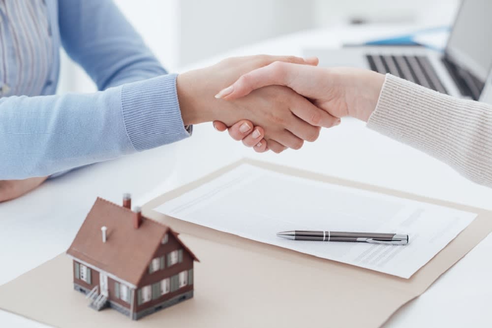 Two Individuals Shaking Hands Over a Paper, Pen and a Tiny House 