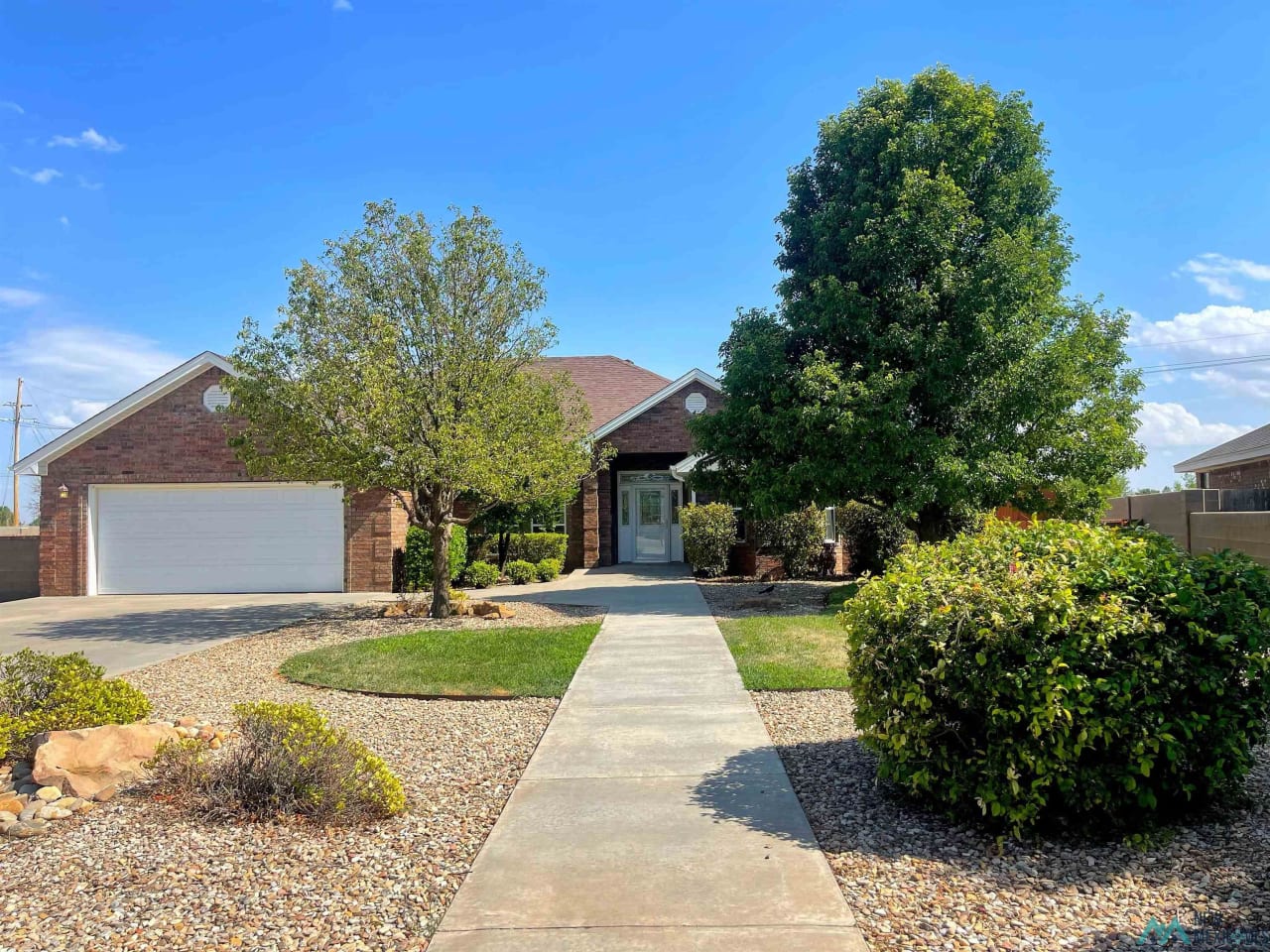 A brick house in a rural area with a sidewalk leading to the house and it’s surrounded by trees and bushes.