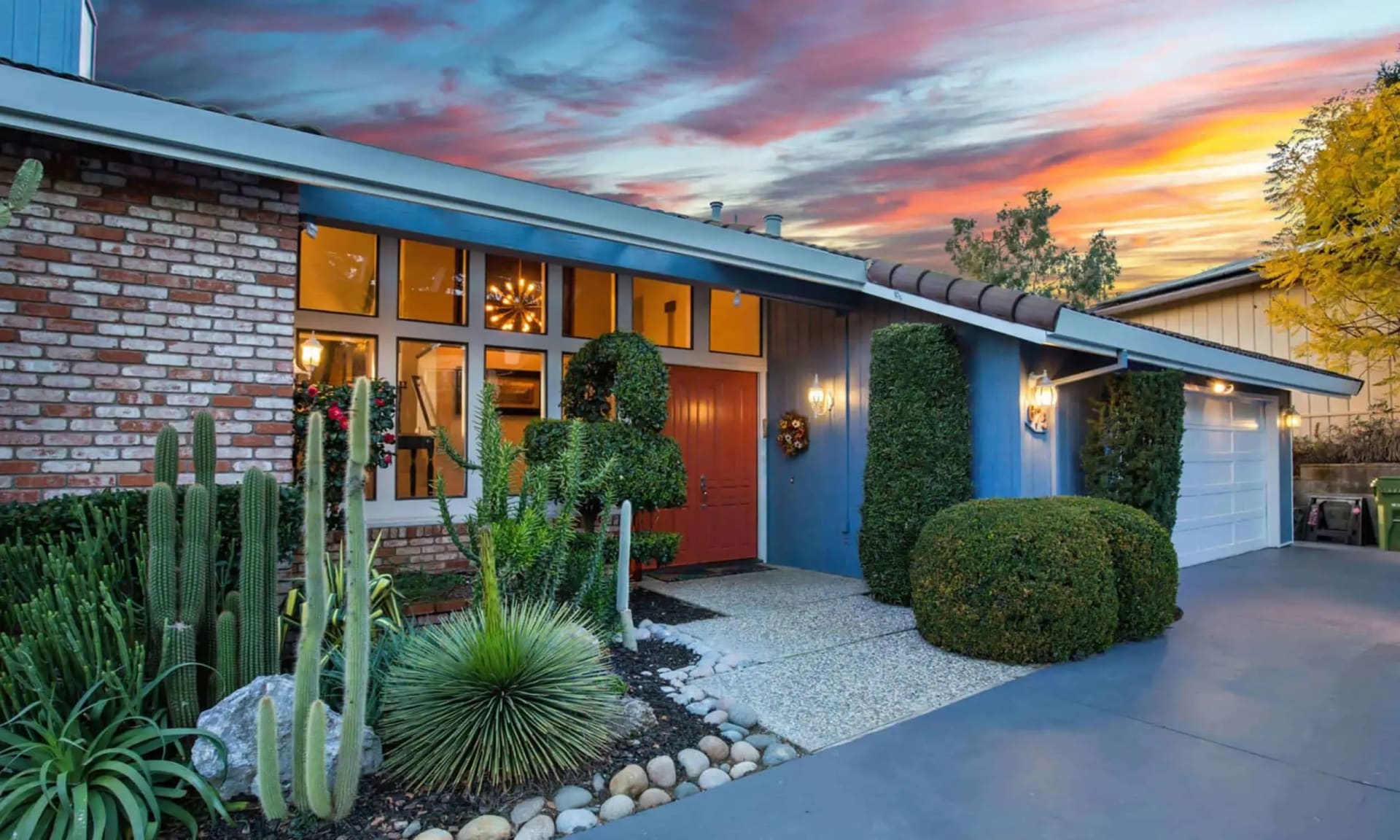 Modern blue-and-brick home with a cactus garden and red front door at sunset.