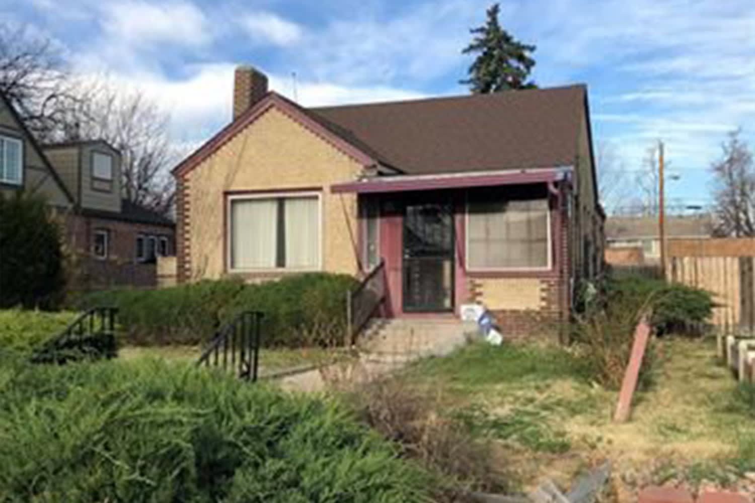 A somewhat dated single-story house with a light brown or tan exterior and a dark brown gabled roof.