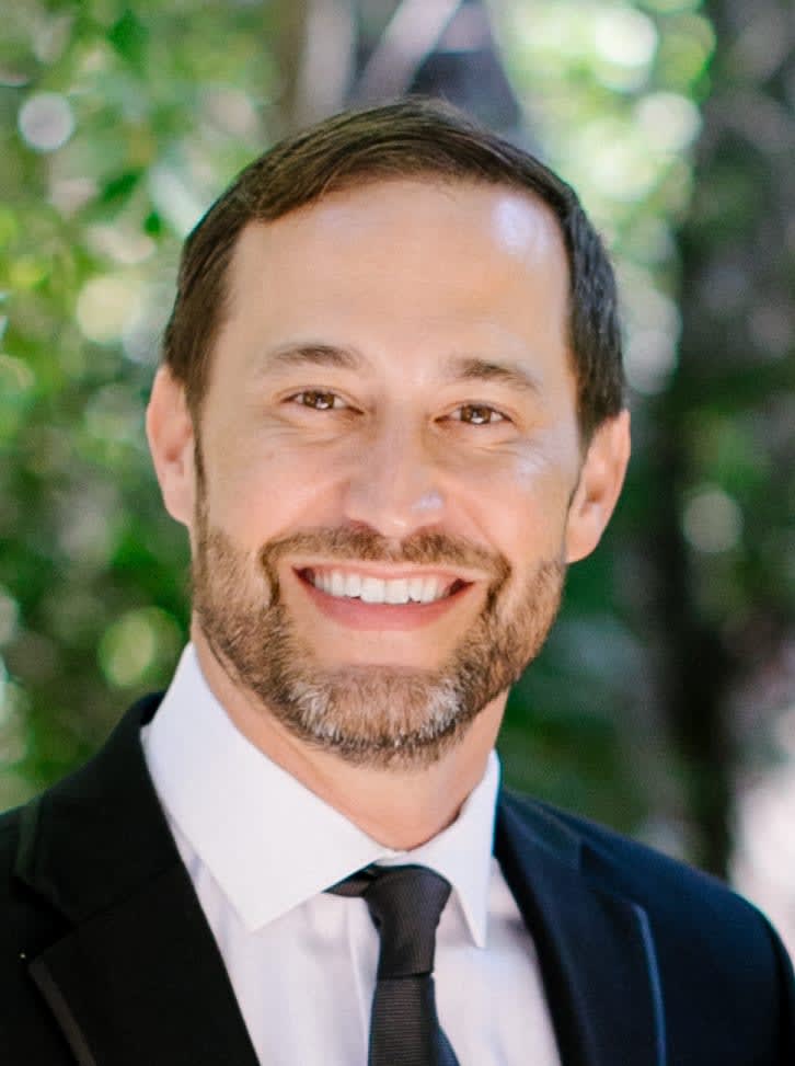 Smiling bearded man in a dark suit and tie against leafy greenery.