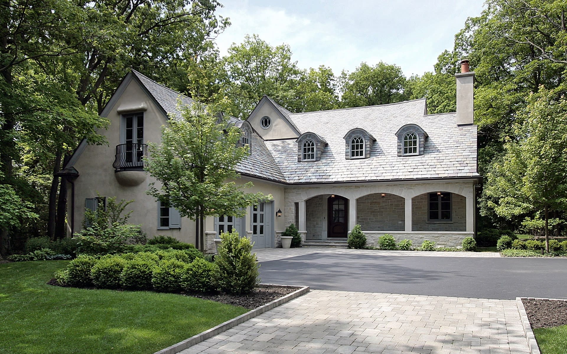 Gray stone-and-stucco house with slate roof, dormers, covered porch, and landscaped driveway.