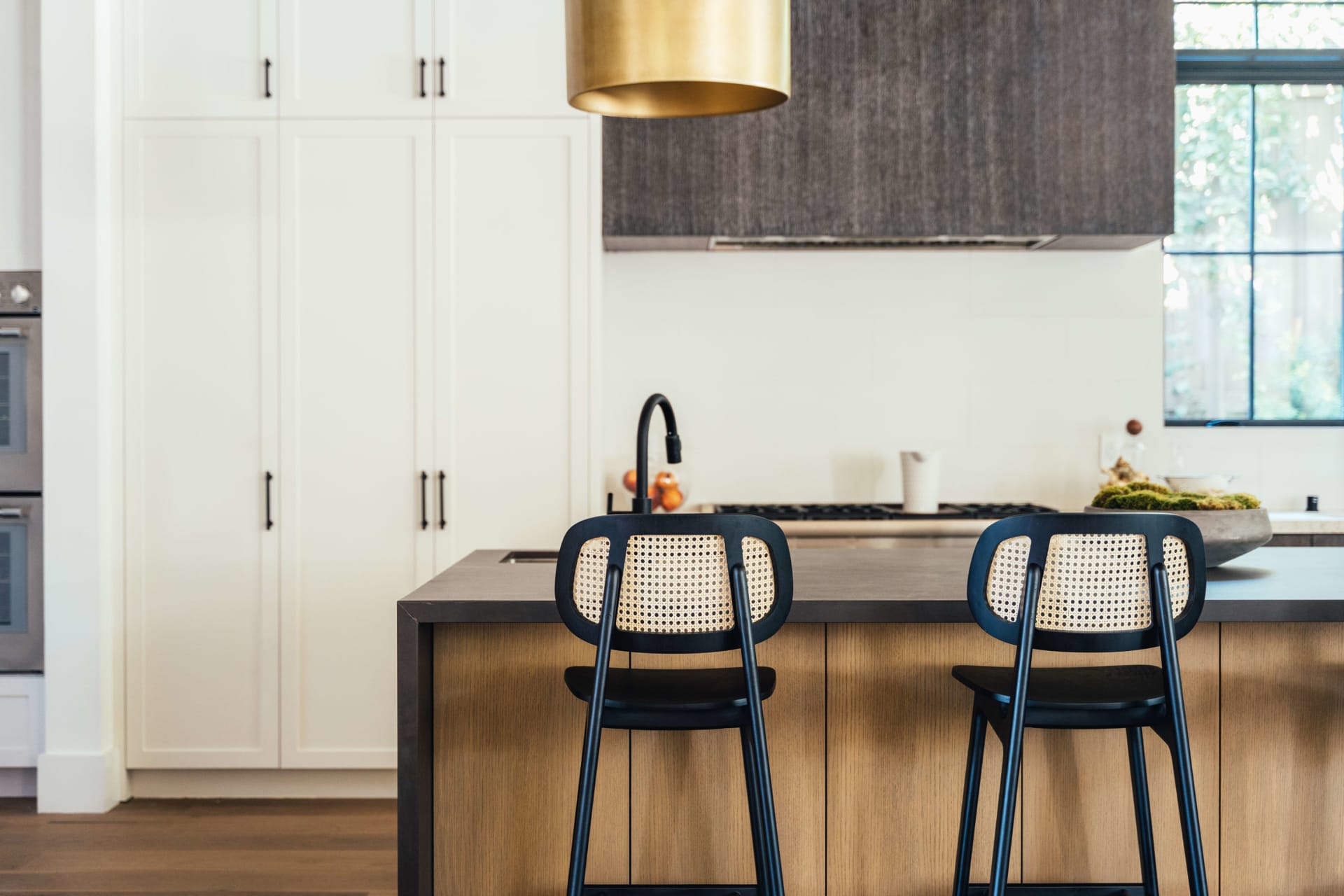  A stylish and modern kitchen features a large island with a dark countertop and light wood paneling, accompanied by two black bar stools with woven cane backs. 