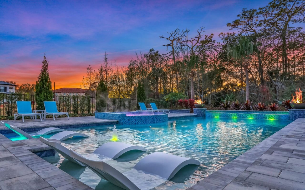 A large, rectangular swimming pool with several in-water chaise lounges and a sun setting on the background.