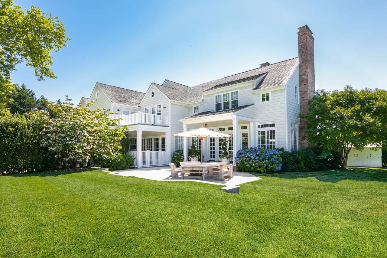 A large white house with a columned porch, a brick walkway, a neat lawn, an outdoor patio, and scattered trees.