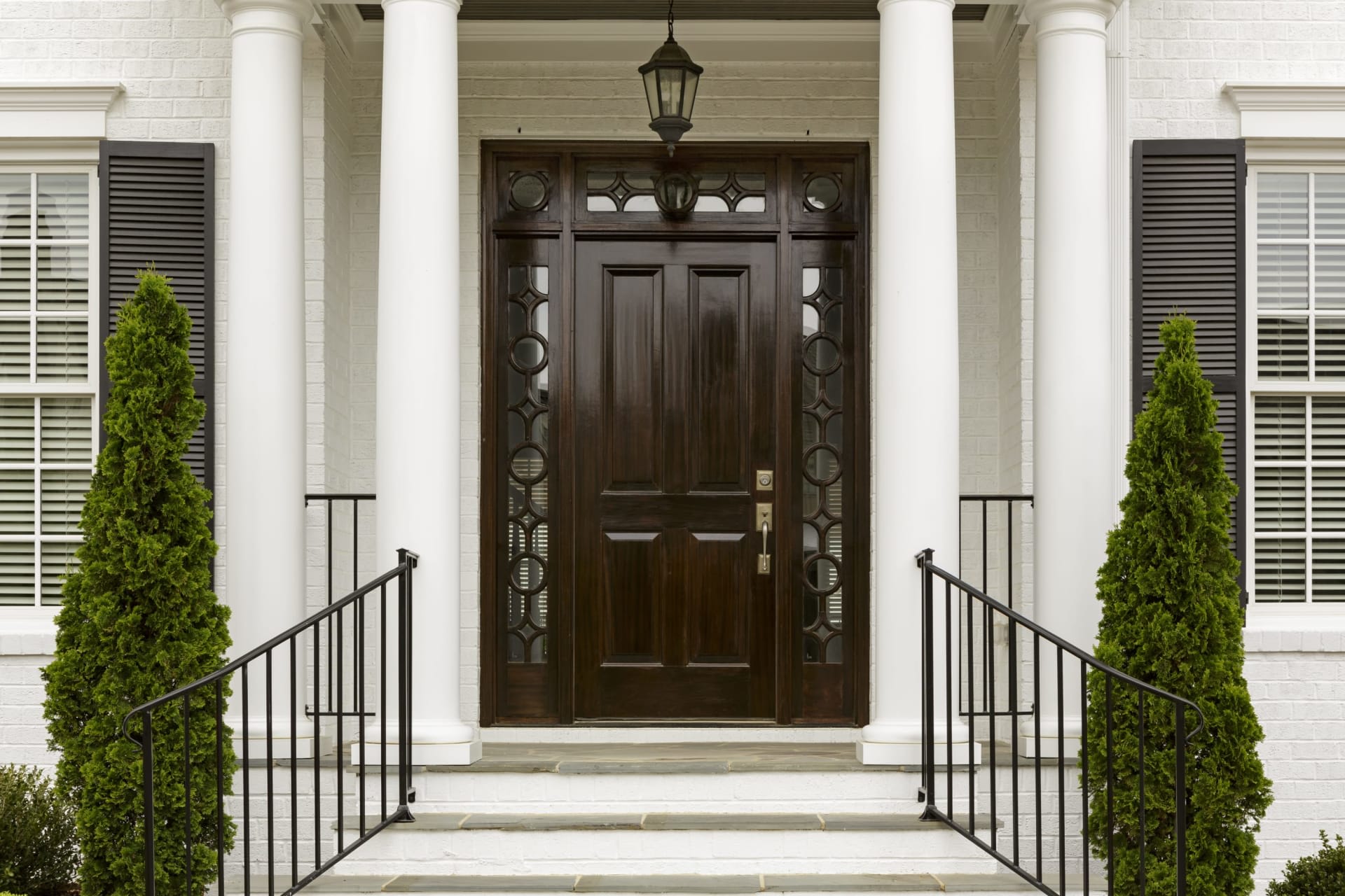 White-brick home entrance with a dark wood door, tall columns, and evergreen shrubs.