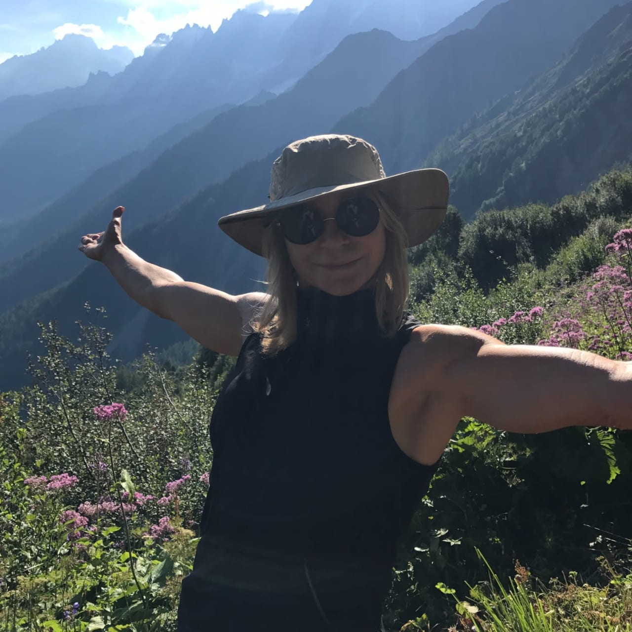 Tracy Curtis standing in the middle of colorful flowers with mountains in the background, wearing a hiking headgear and sunglasses.