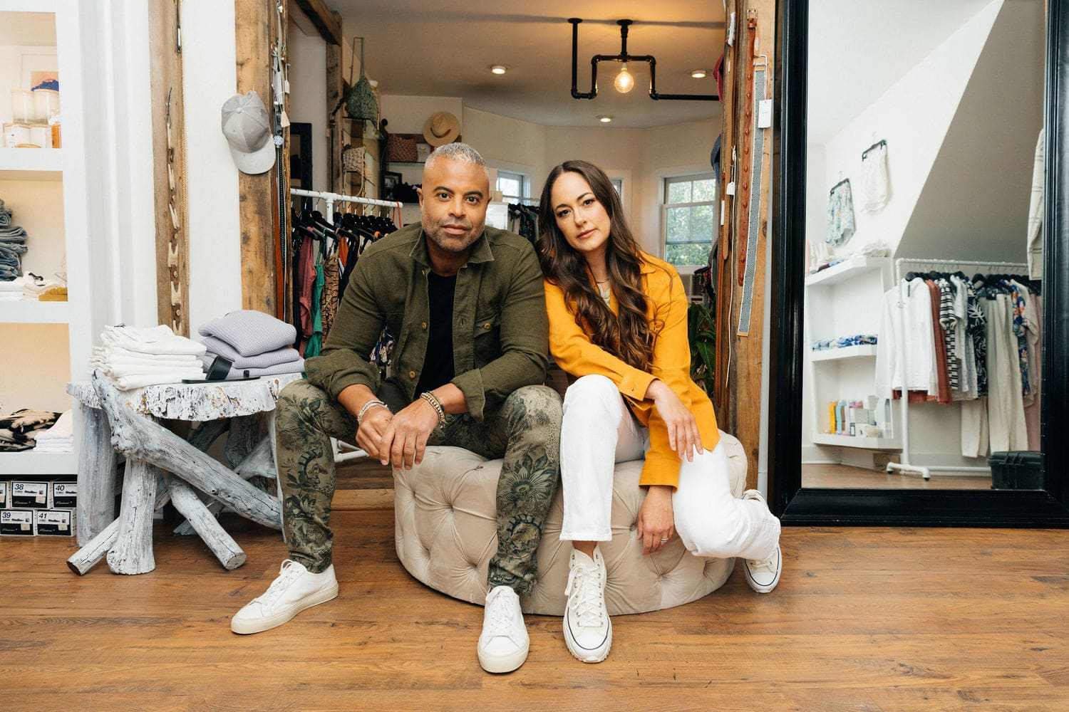 A man and woman seated in a clothing boutique, surrounded by clothing racks and folded garments.