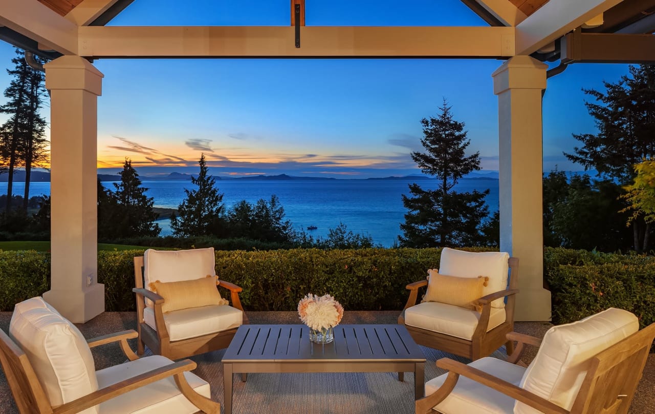 Outdoor patio with cushioned chairs and a table overlooking a serene ocean view at sunset, framed by trees and a garden.