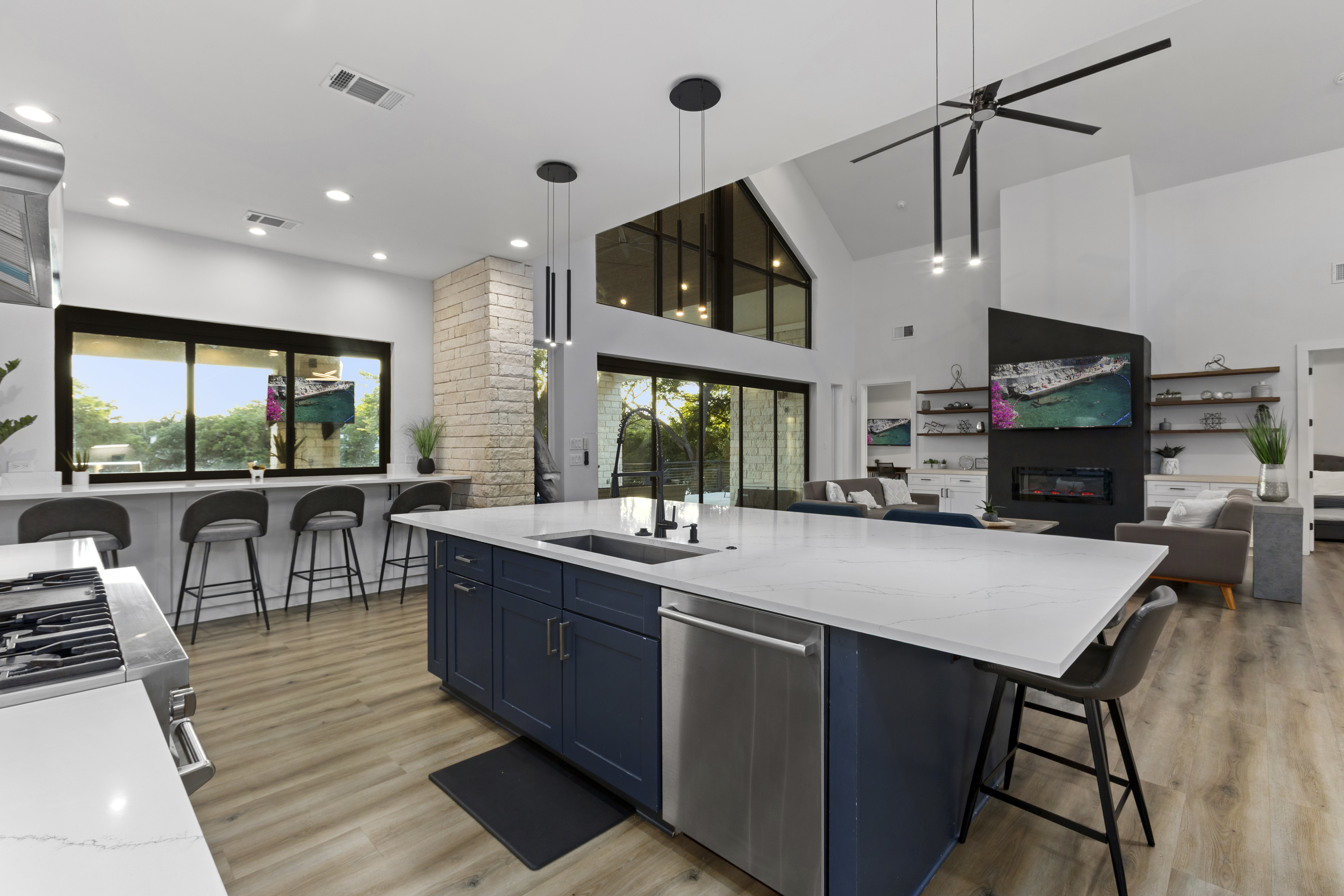 Kitchen island with a white marble-look top over navy cabinets, a black faucet and stainless dishwasher, looking past a breakfast bar window and black pendant lights to the vaulted great room with a fireplace wall