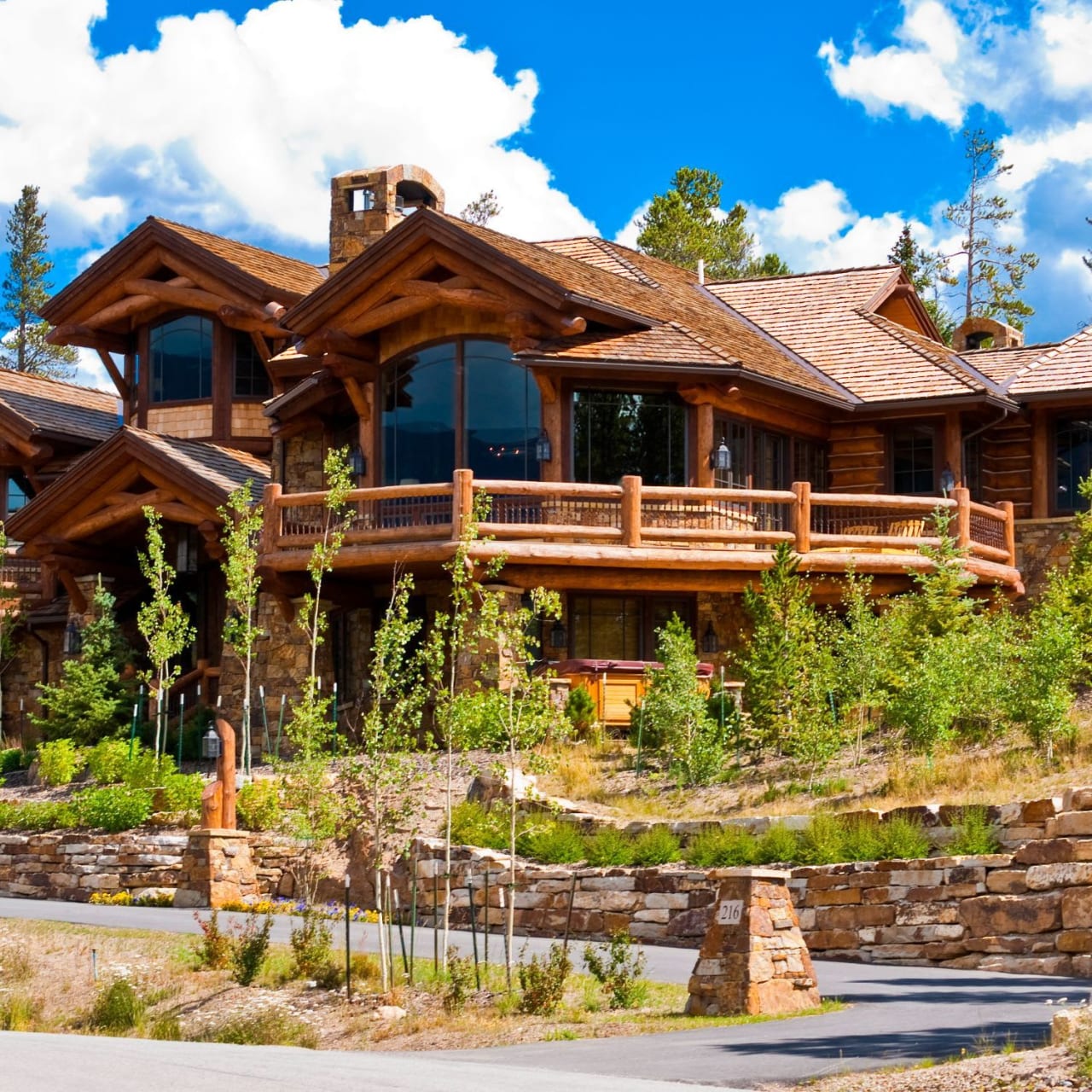 A large log home with a big deck, sitting on a hilltop surrounded by trees.