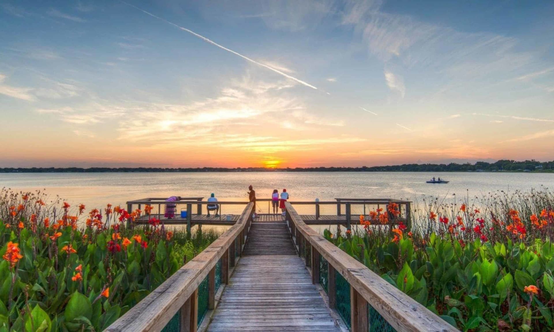 Waterfront boardwalk lined with orange flowers, with people watching the sunset over the water.