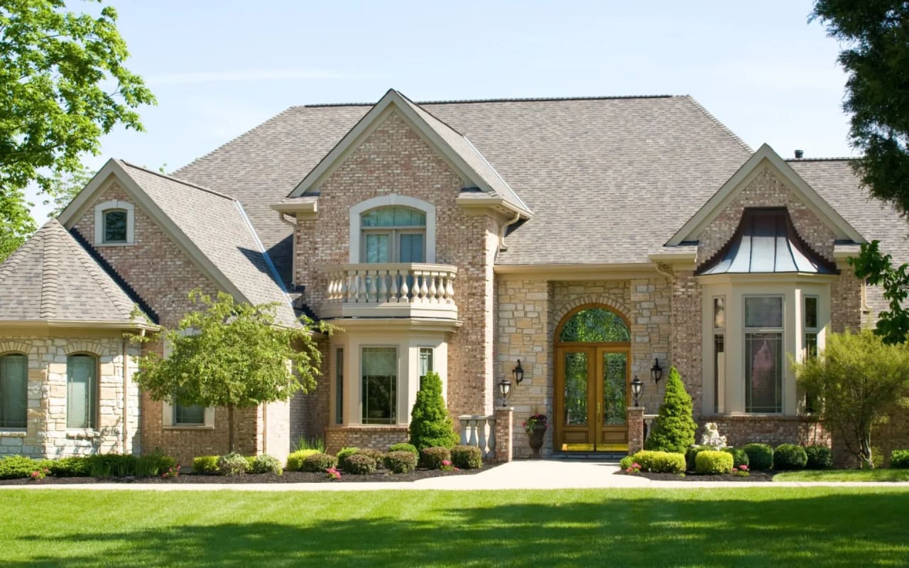 A Colonial-style, two-story brick house with a wraparound porch, a balustrade, front lawn, and sidewalks on both sides.