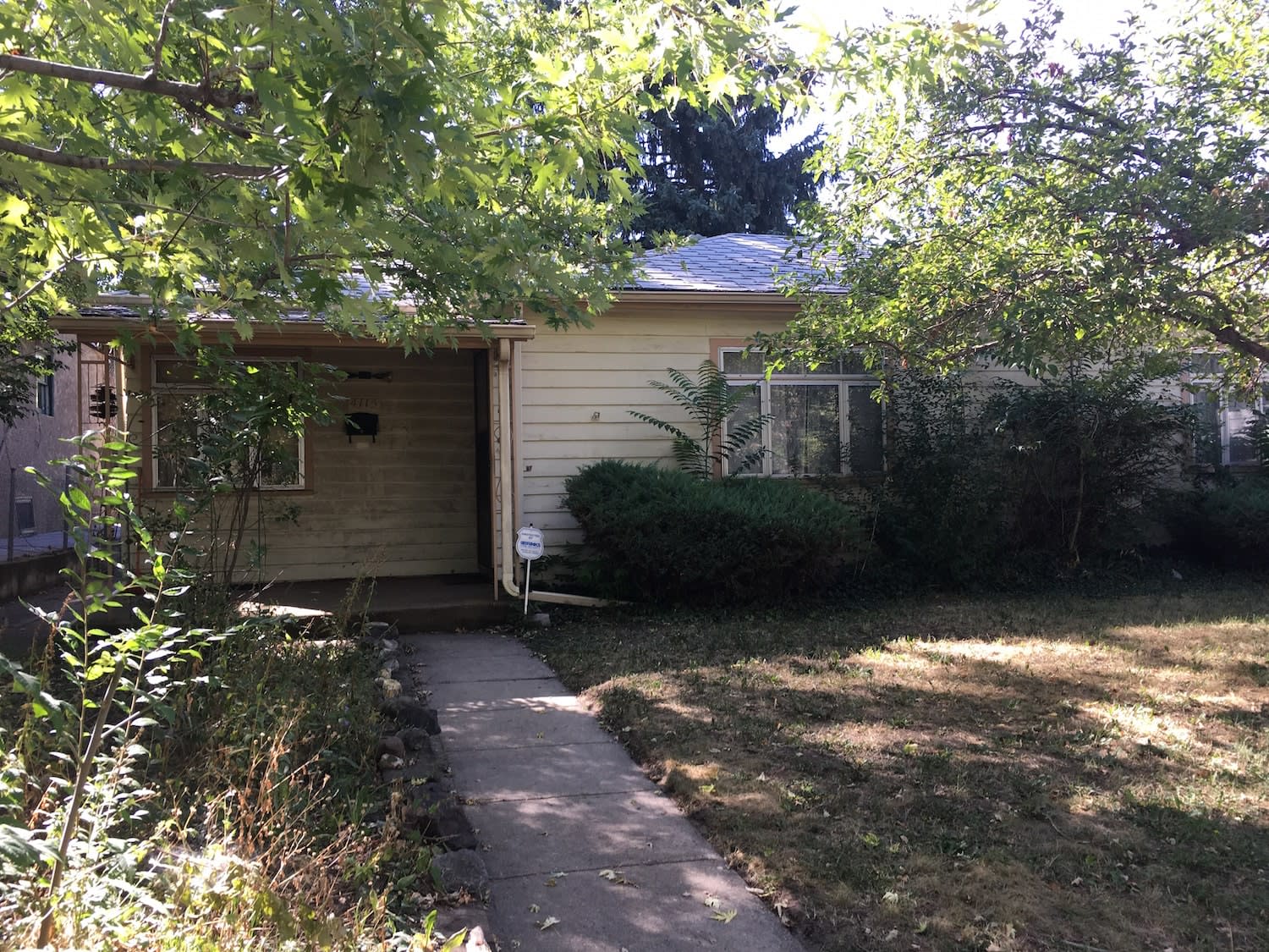  A single-story, light-colored house with a covered porch on the left, partially obscured by lush green trees and overgrown bushes.