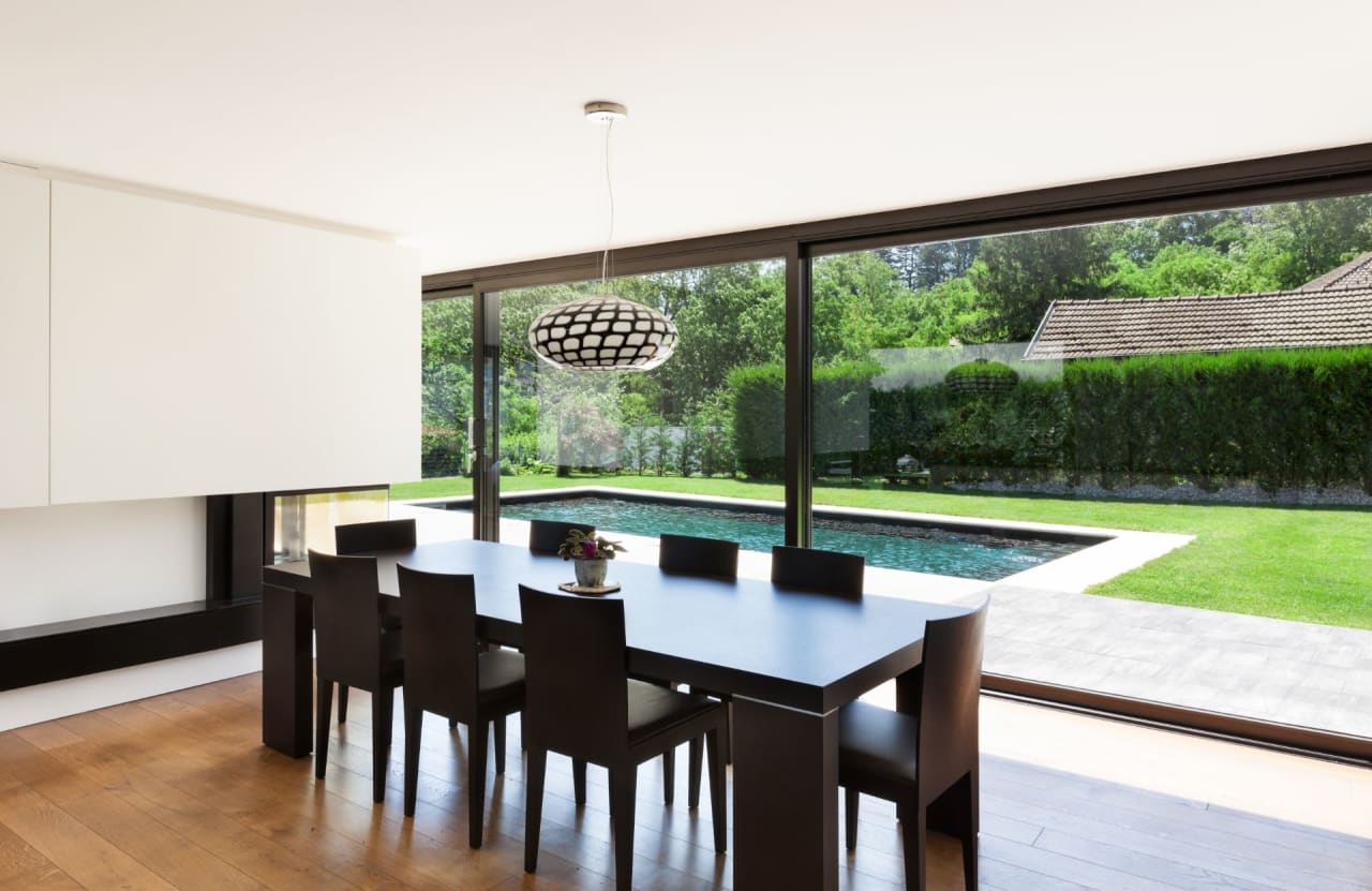 A modern dining room with a modern table and chairs. There is a swimming pool visible through the windows behind the table.