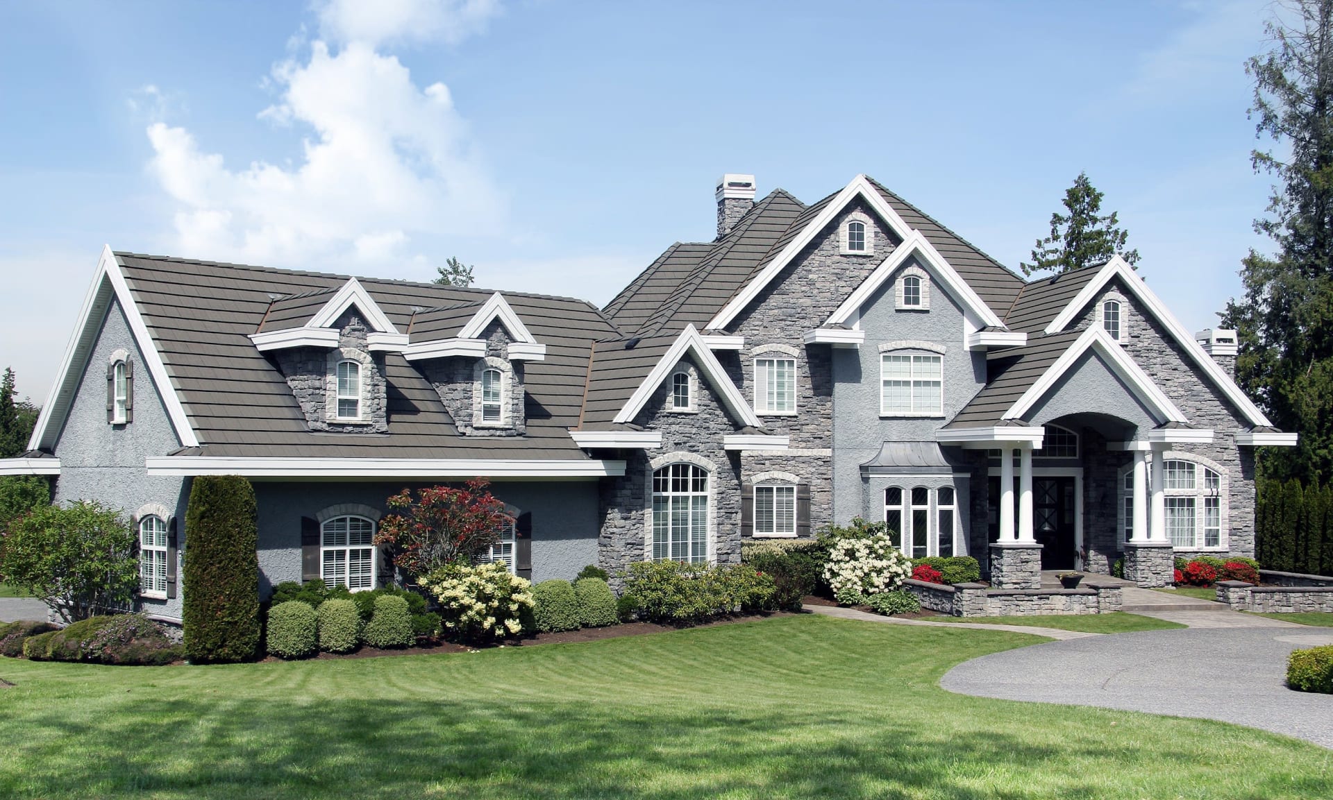 Large gray stone-accented house with multiple gables, landscaped lawn, and curved driveway.