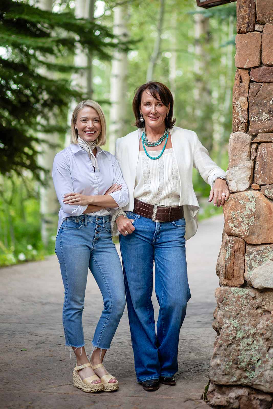Two women pose together beside a stone wall on a leafy path.