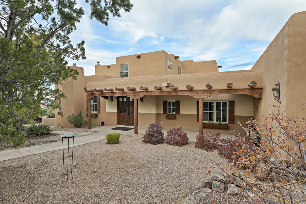 The front of a large adobe house with a covered porch in a desert landscape.