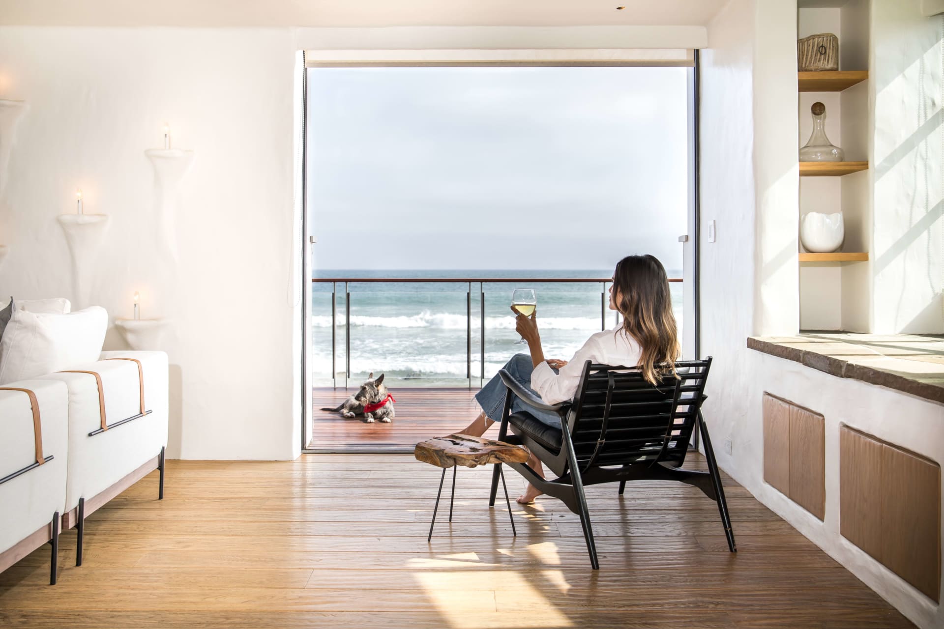 A person with long, dark hair sits facing the ocean in a black modern chair on a wooden deck, holding a glass of white wine. 