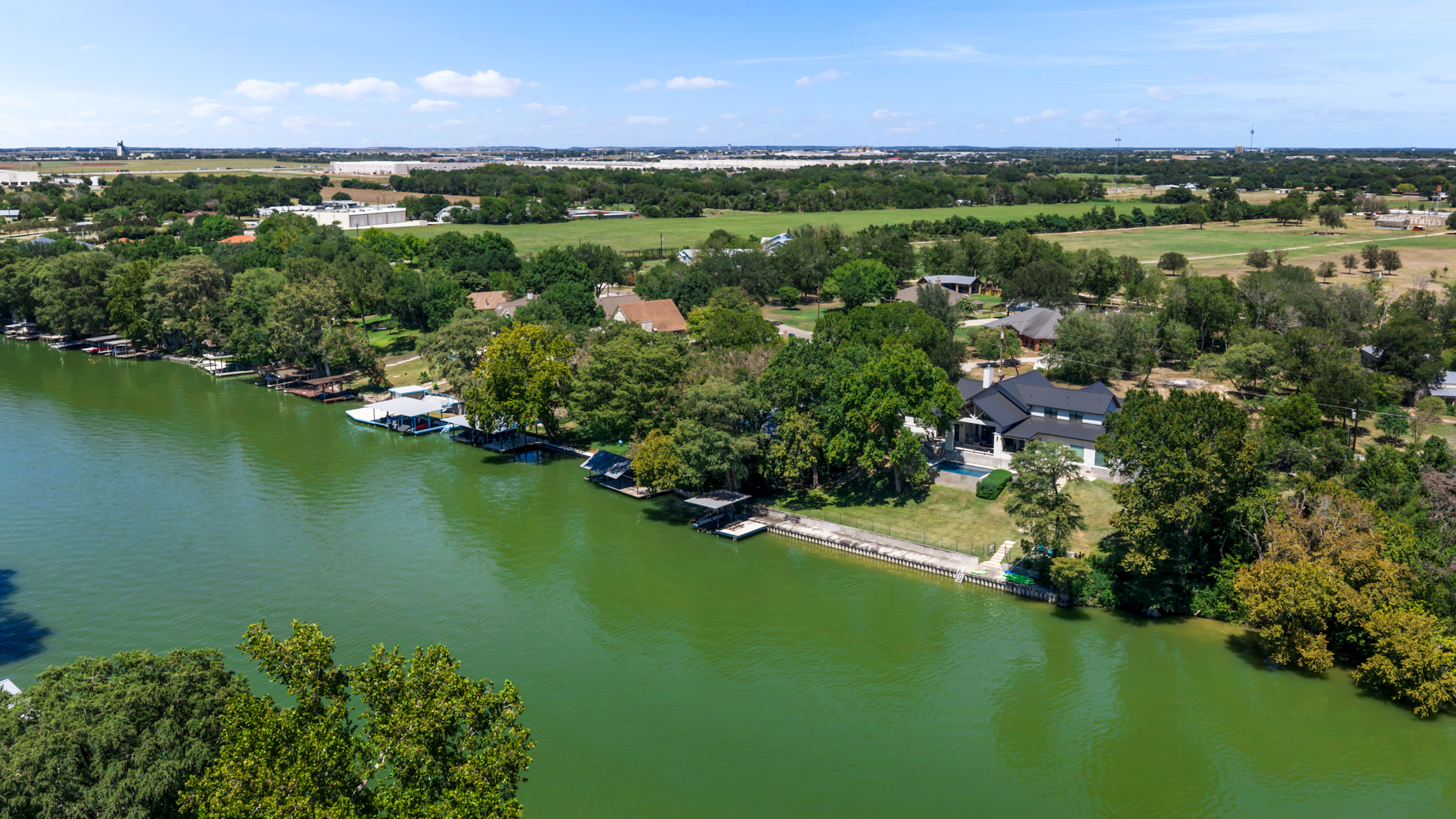 Wide aerial view of Lake Placid in Seguin with green water, boathouses and docks along a tree-lined shoreline, a two-story home with a pool near the center, and open fields beyond