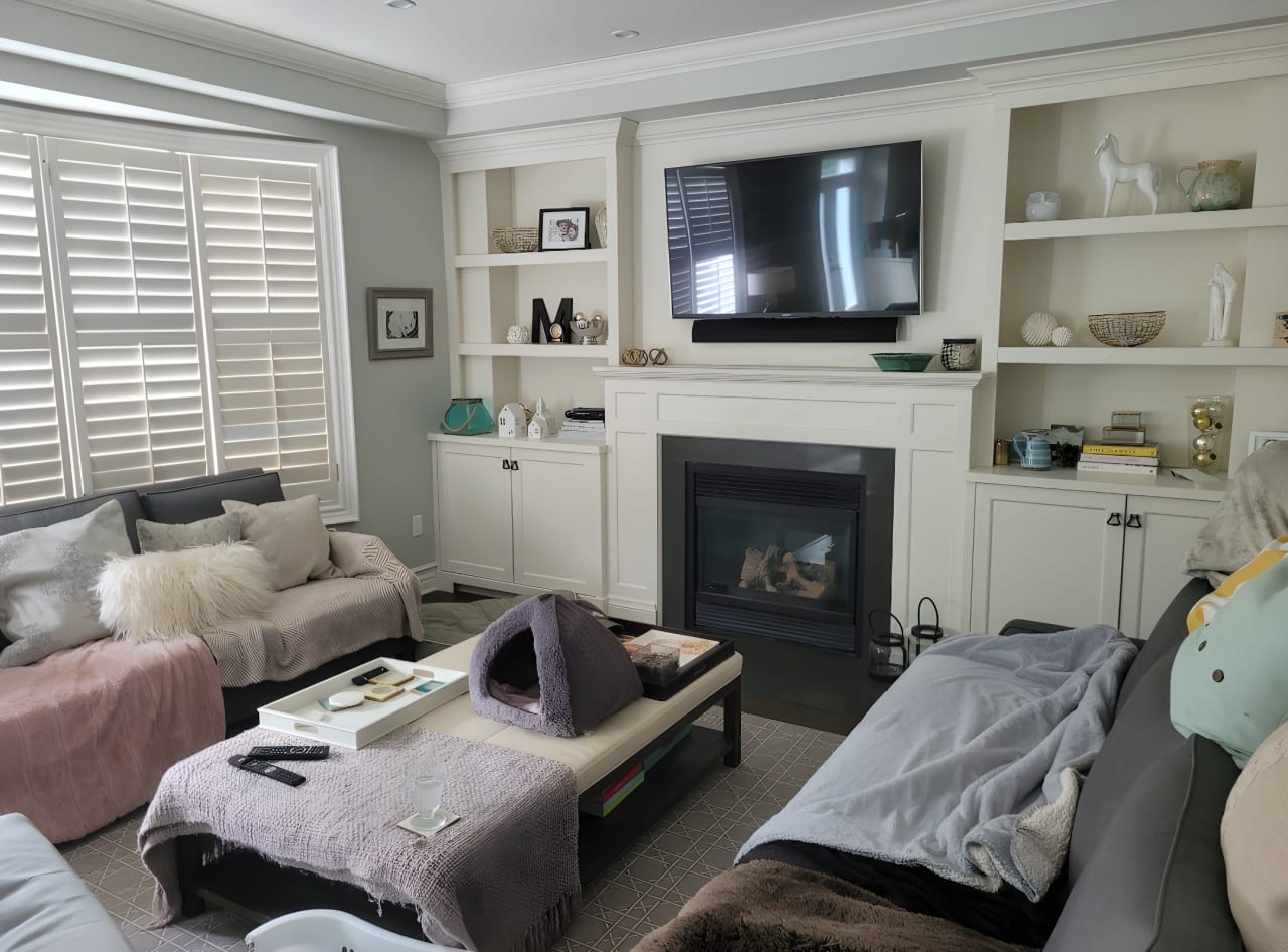 A cluttered living room with a fireplace, two couches, a coffee table, and a flat-screen TV mounted on the wall.