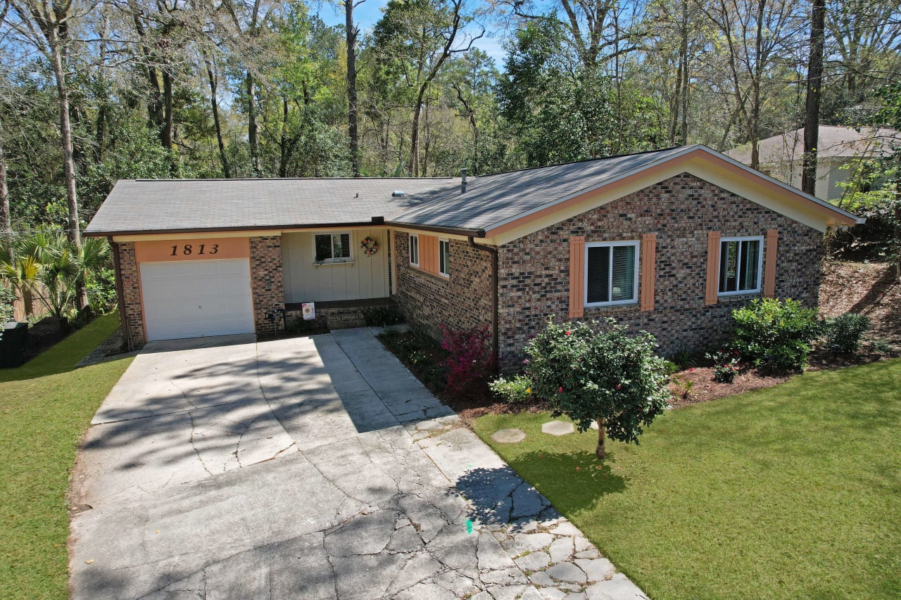 An aerial view of a single-story house with a garage, driveway, and front yard. The house has a mix of brick and siding on the exterior.